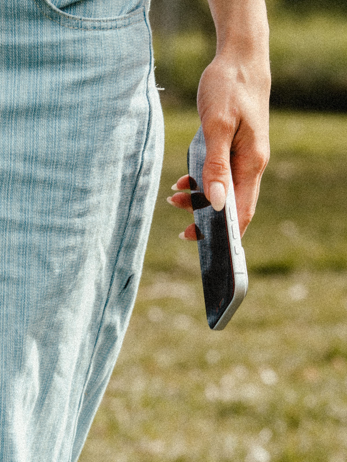 A close-up lifestyle shot of a person wearing light blue denim jeans, holding a black smartphone in their hand against a blurred grassy outdoor background.