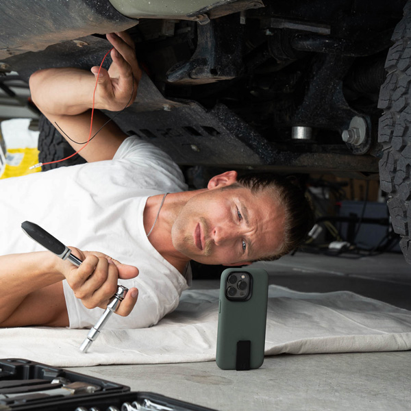 A man is laying down on the ground working on car and using a Motus phone case. The phone case is standing up on its own due to the quick draw clip.